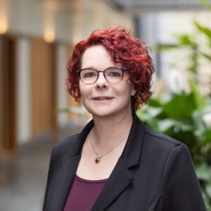 Mitarbeiterin der GfG mit Brille und schwarzem Blazer im hellen Flur