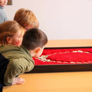 Drei Kinder betrachten neugierig eine Amtskette in einer roten Schaukassette