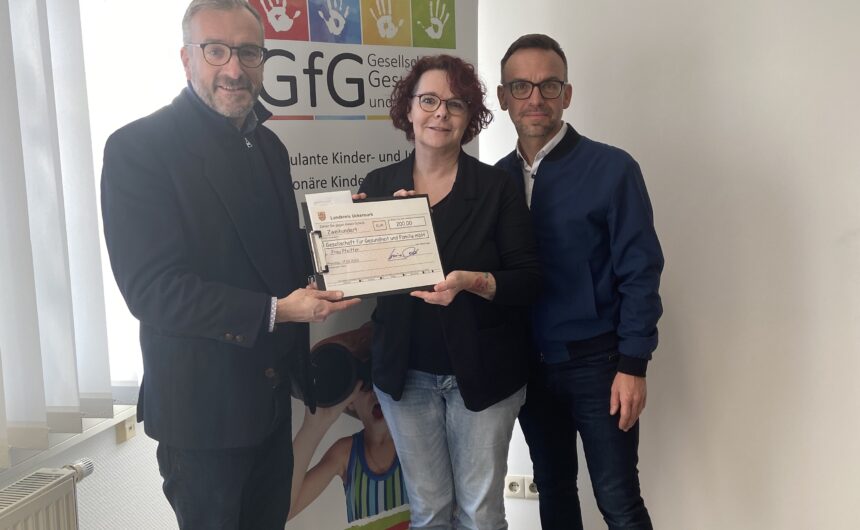 Spendenübergabe_GfG_19022026_Foto_Sandra_Kobelt (1) Scheckübergabe vor dem Rollup der GfG Gesellschaft für Gesundheit und Familie – drei Personen halten gemeinsam einen Spendenscheck