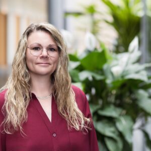 Mitarbeiterin der GfG mit Brille und bordeauxrotem Pullover im hellen Flur